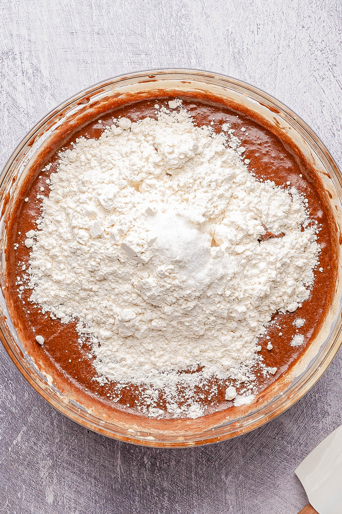 A glass bowl filled with chocolate batter topped with a mound of flour, ready to be mixed, sits on a light textured surface.