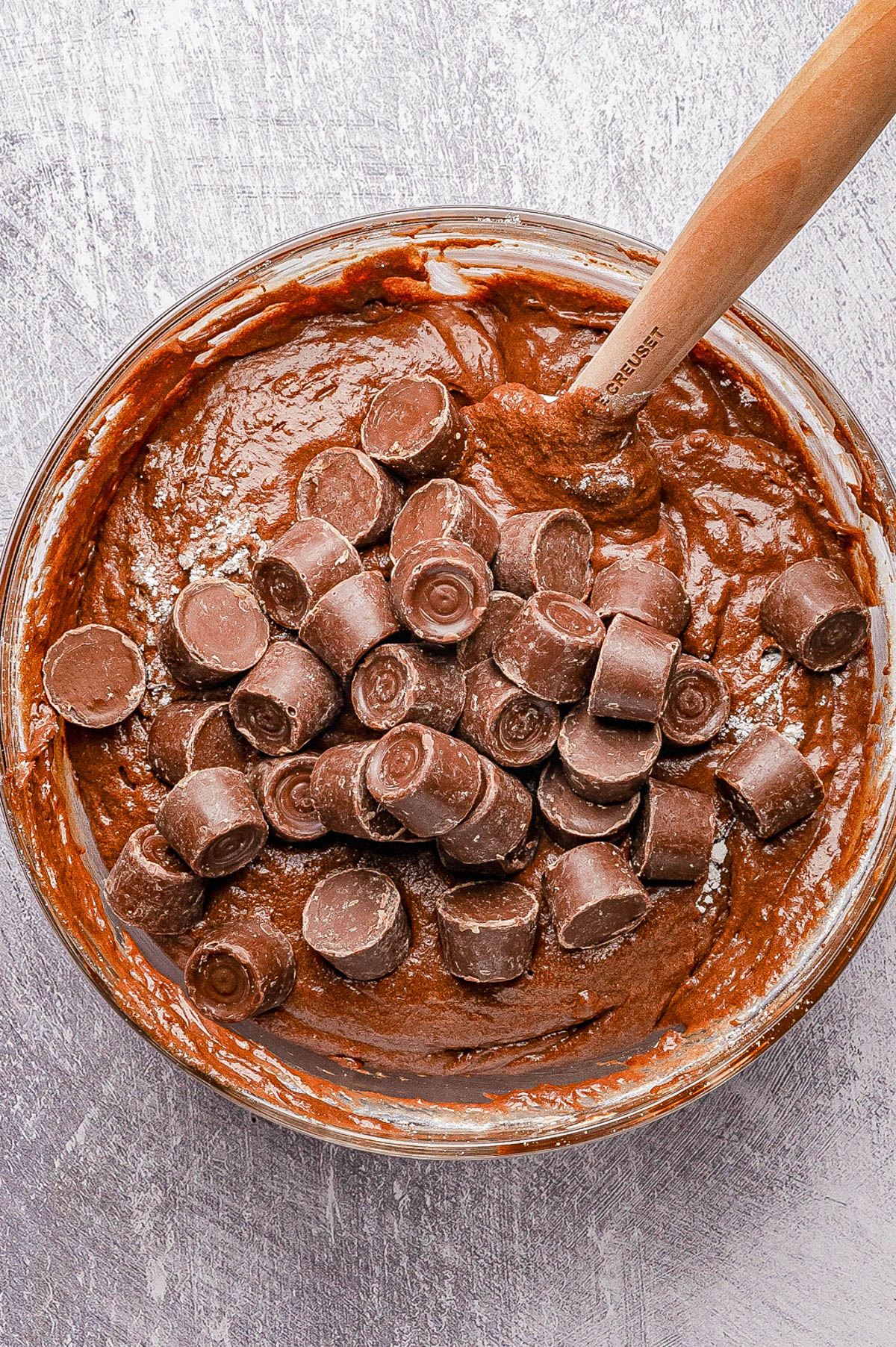A glass bowl filled with chocolate batter and topped with unwrapped chocolate candies, with a wooden spoon resting inside.