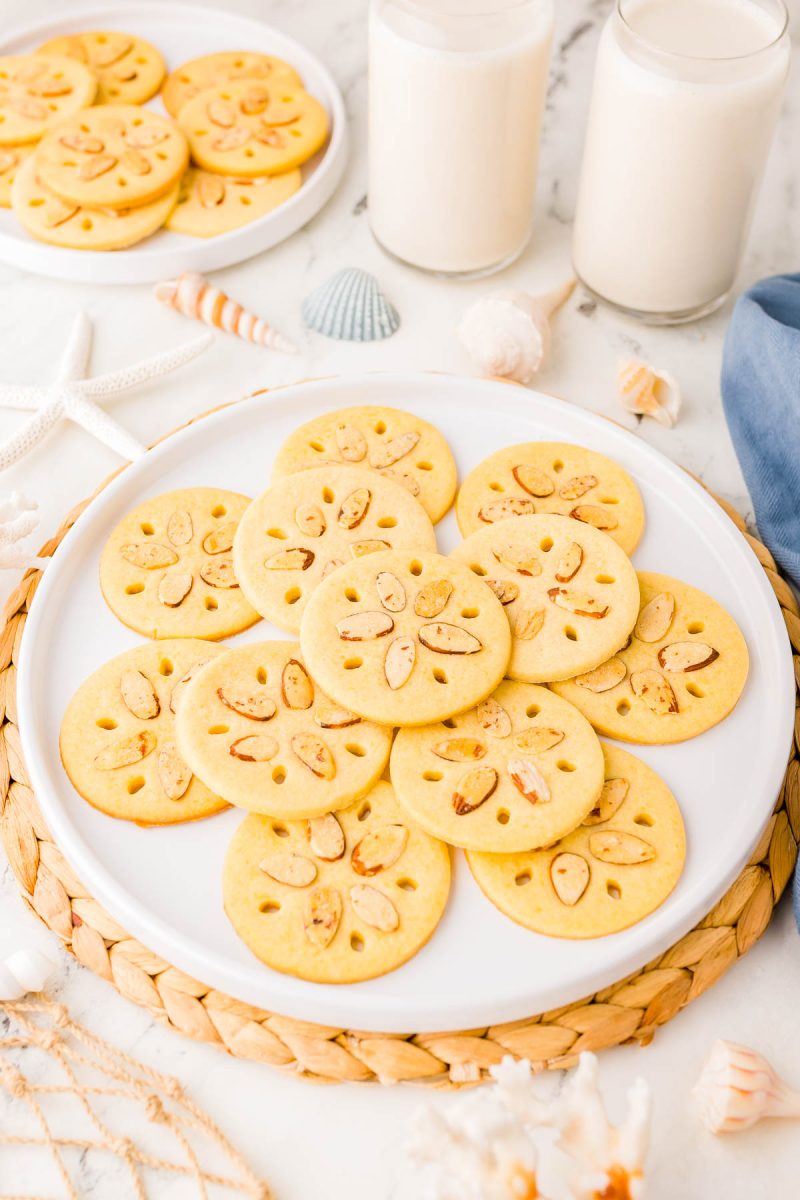 A plate of round cookies with almond slices on top, next to two glasses of milk and seashell decorations on a white surface.