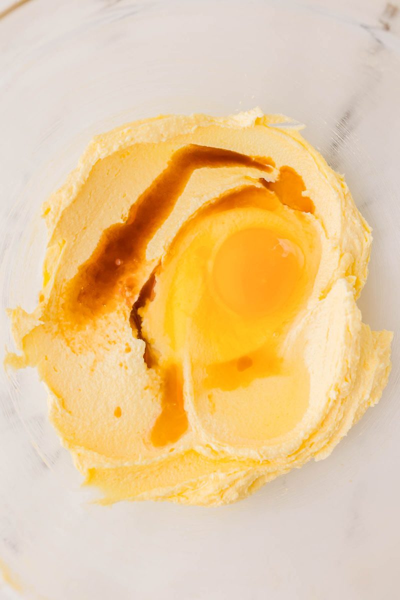 A glass bowl containing creamed butter and sugar, with an egg and vanilla extract added on top, ready to be mixed.