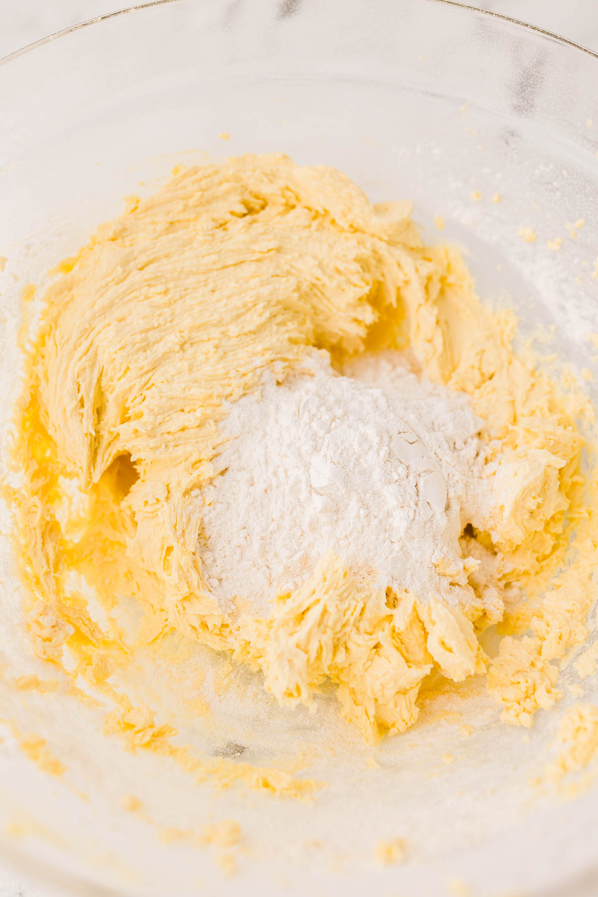 A glass mixing bowl containing creamed butter and sugar with flour added on top, ready to be mixed.