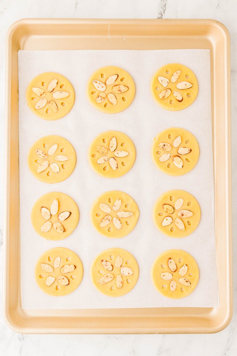 A baking sheet with twelve round, unbaked cookies topped with almond slices arranged in a flower pattern on parchment paper.