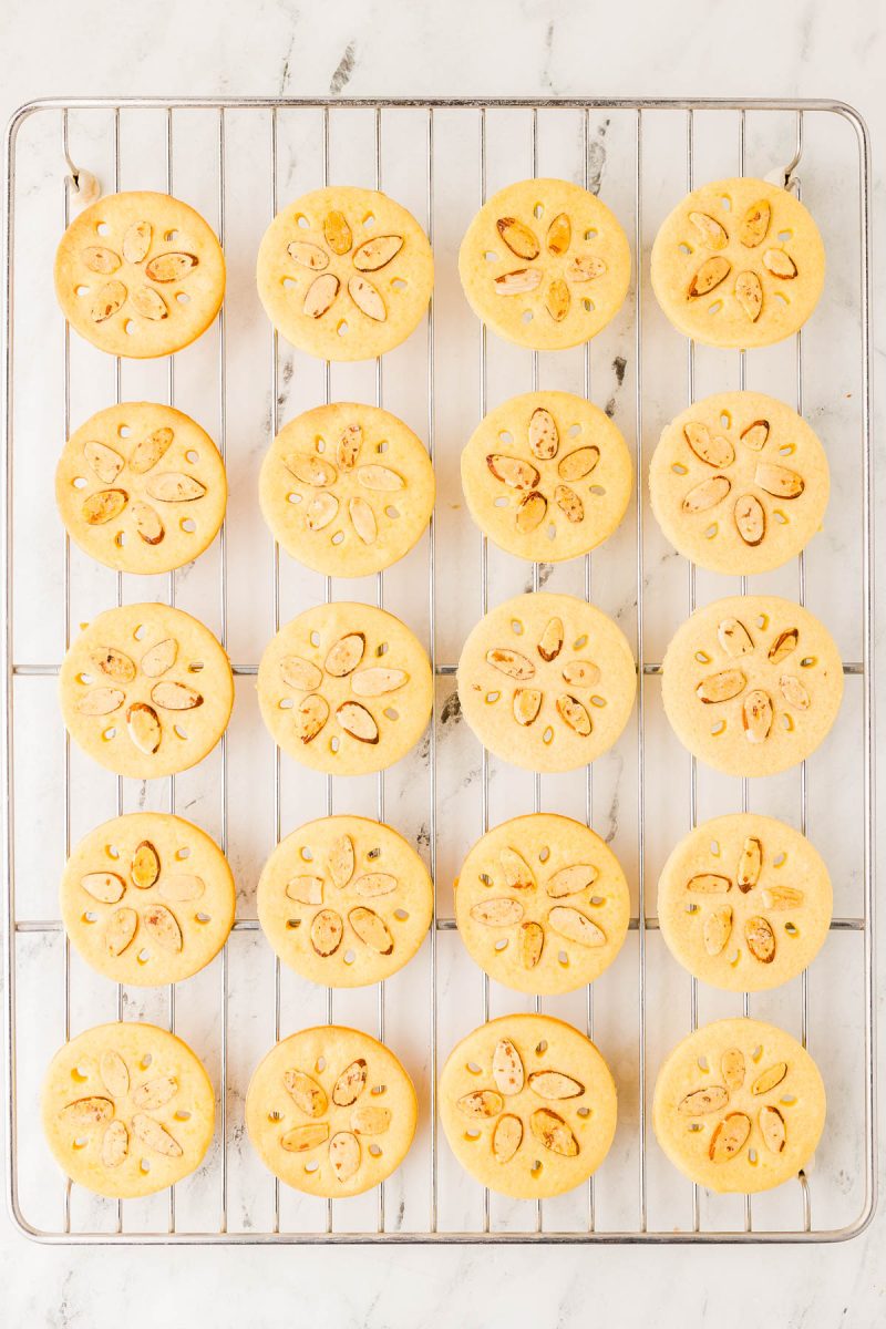 Rows of round cookies with sliced almonds arranged in a flower pattern on a cooling rack over a white marble surface.