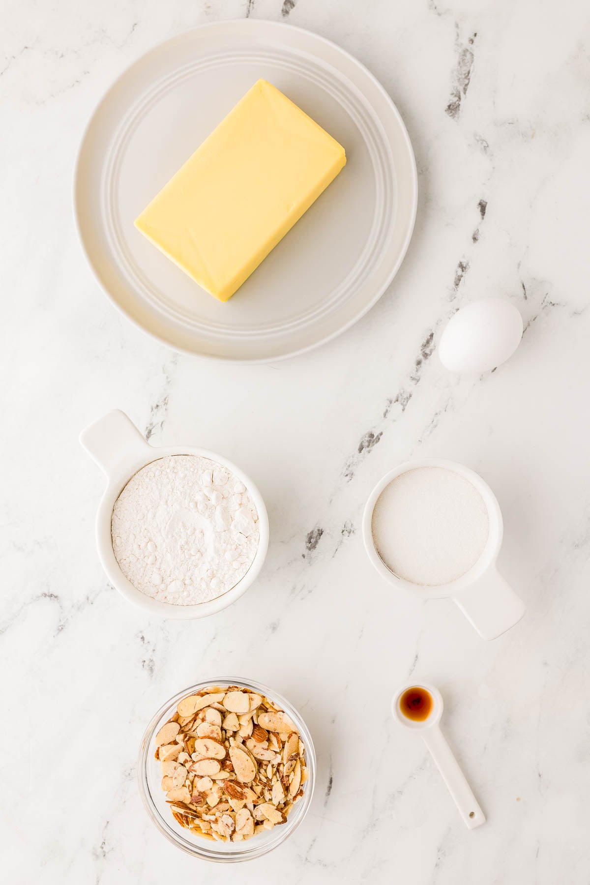 Butter on a plate, an egg, flour, sugar, chopped nuts in a bowl, and a spoon of vanilla extract arranged on a marble countertop.