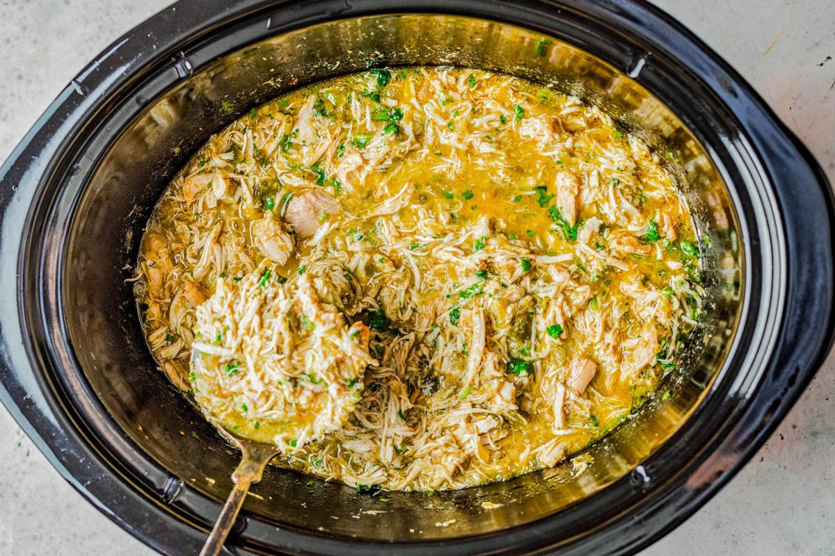 Shredded chicken in a thick, yellow broth with herbs in a black slow cooker, with a ladle lifting some of the mixture.