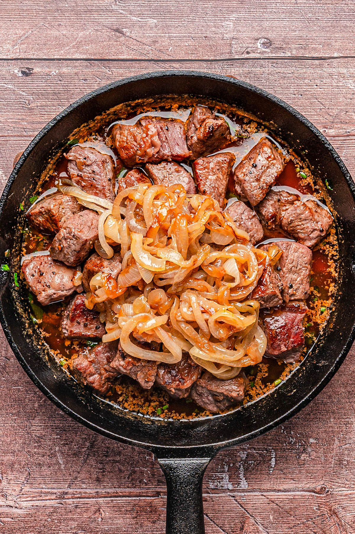 A cast iron skillet filled with cooked beef cubes topped with sautรฉed onions, set on a wooden surface.