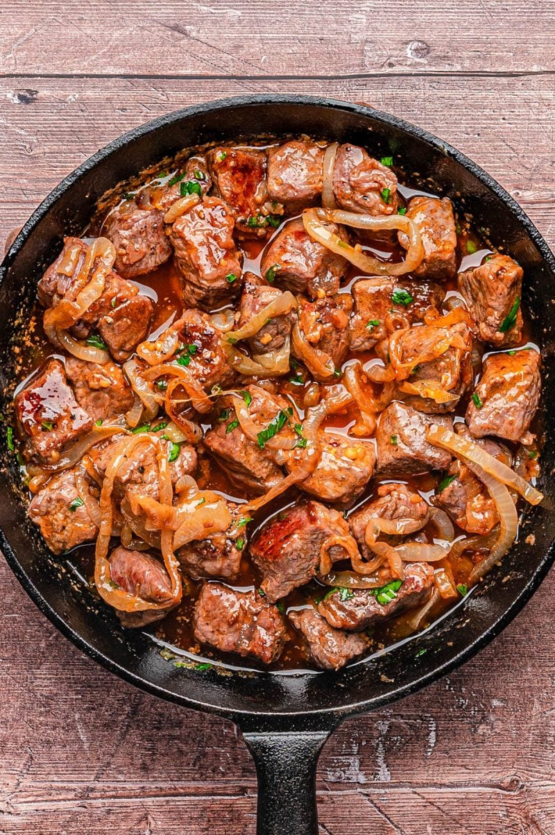 A cast iron skillet filled with cooked beef cubes, sautéed onions, fresh herbs, and sauce, placed on a wooden surface.