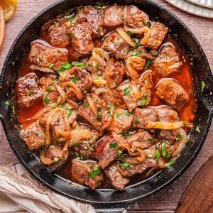 A cast iron skillet filled with cooked beef cubes, sliced onions, and chopped herbs in a brown sauce, placed on a wooden table with plates and utensils nearby.