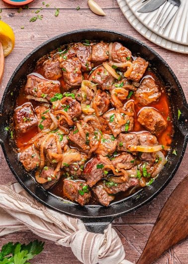 A cast iron skillet filled with cooked beef cubes, sliced onions, and chopped herbs in a brown sauce, placed on a wooden table with plates and utensils nearby.