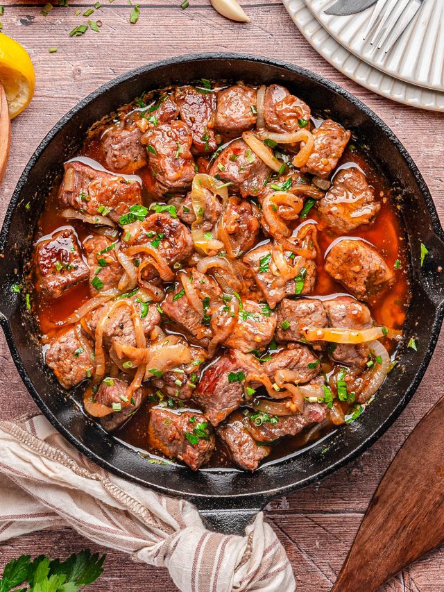 A cast iron skillet filled with cooked beef cubes, sliced onions, and chopped herbs in a brown sauce, placed on a wooden table with plates and utensils nearby.