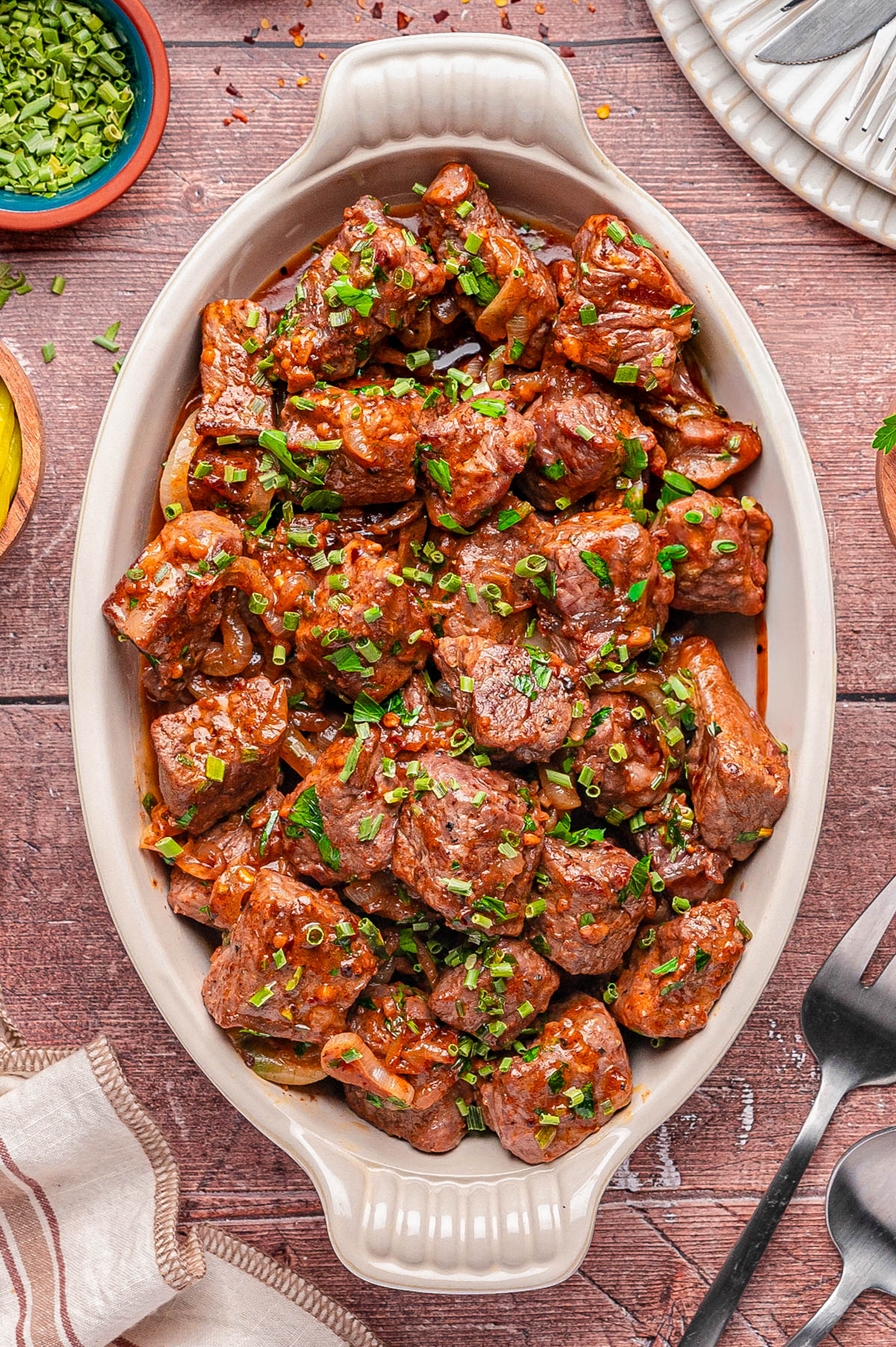 A white oval dish filled with cooked beef tips garnished with chopped green herbs, placed on a wooden table with utensils and small bowls nearby.