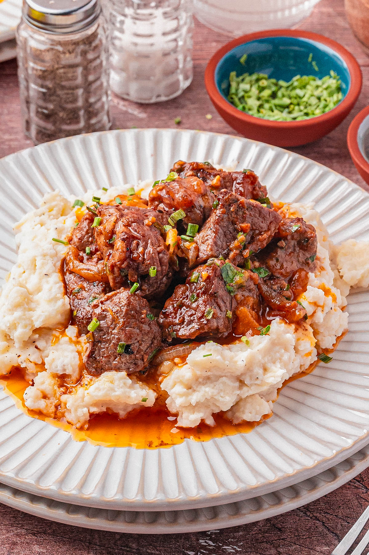 A plate of mashed potatoes topped with braised beef in a red sauce, garnished with chopped chives, on a white textured plate.