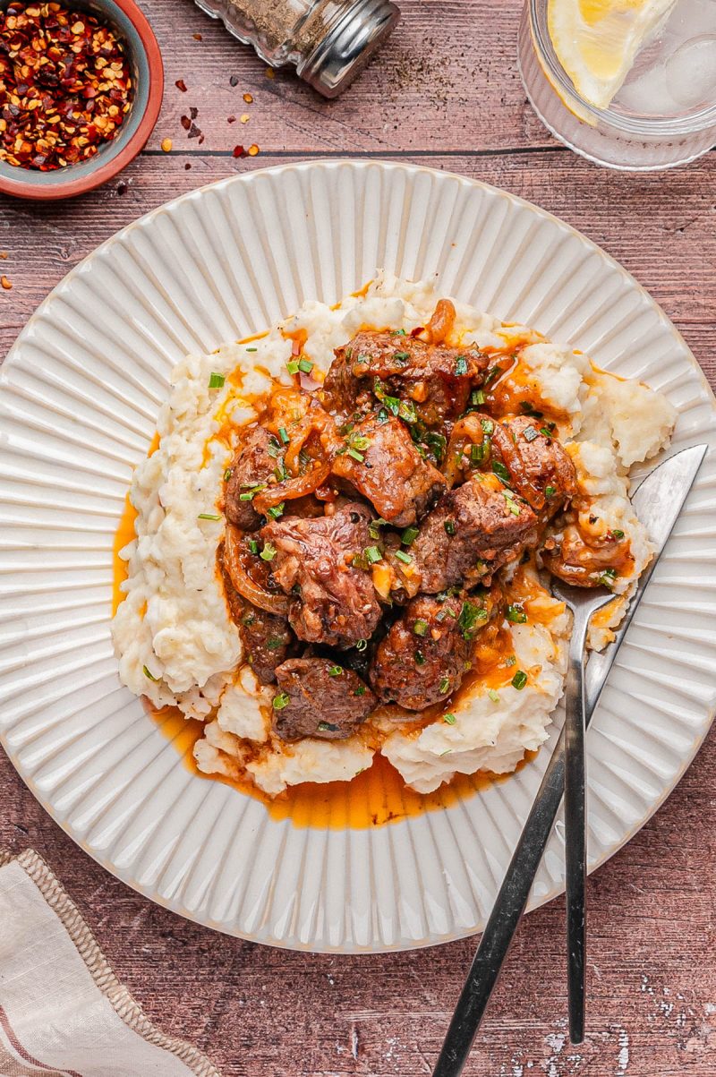A plate of mashed potatoes topped with a saucy beef stew, garnished with herbs, served with a fork and knife on a white plate.