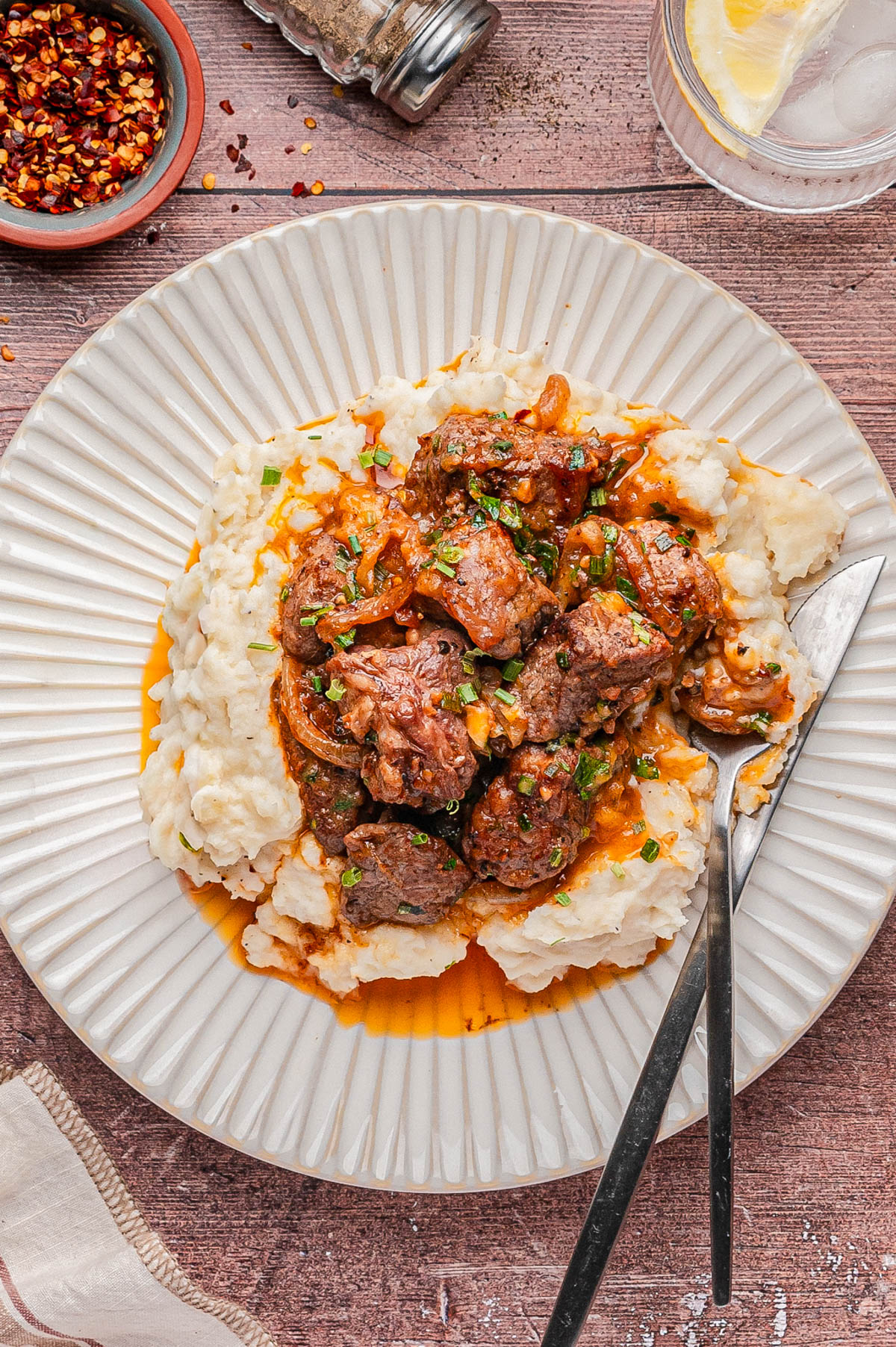A plate of mashed potatoes topped with a saucy beef stew, garnished with herbs, served with a fork and knife on a white plate.