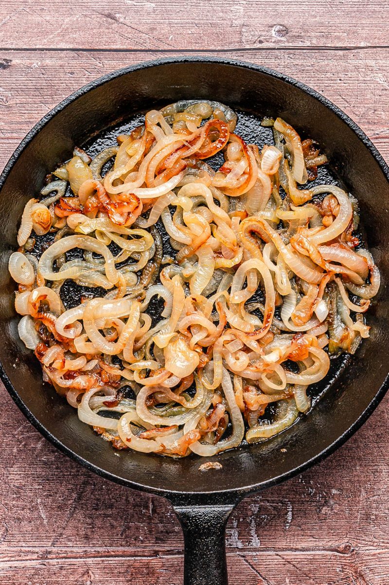 Sliced onions being caramelized in a black cast iron skillet on a wooden surface.