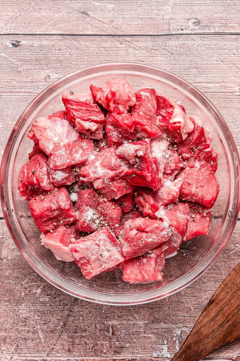 A glass bowl containing raw beef cubes seasoned with salt and pepper, placed on a wooden surface.