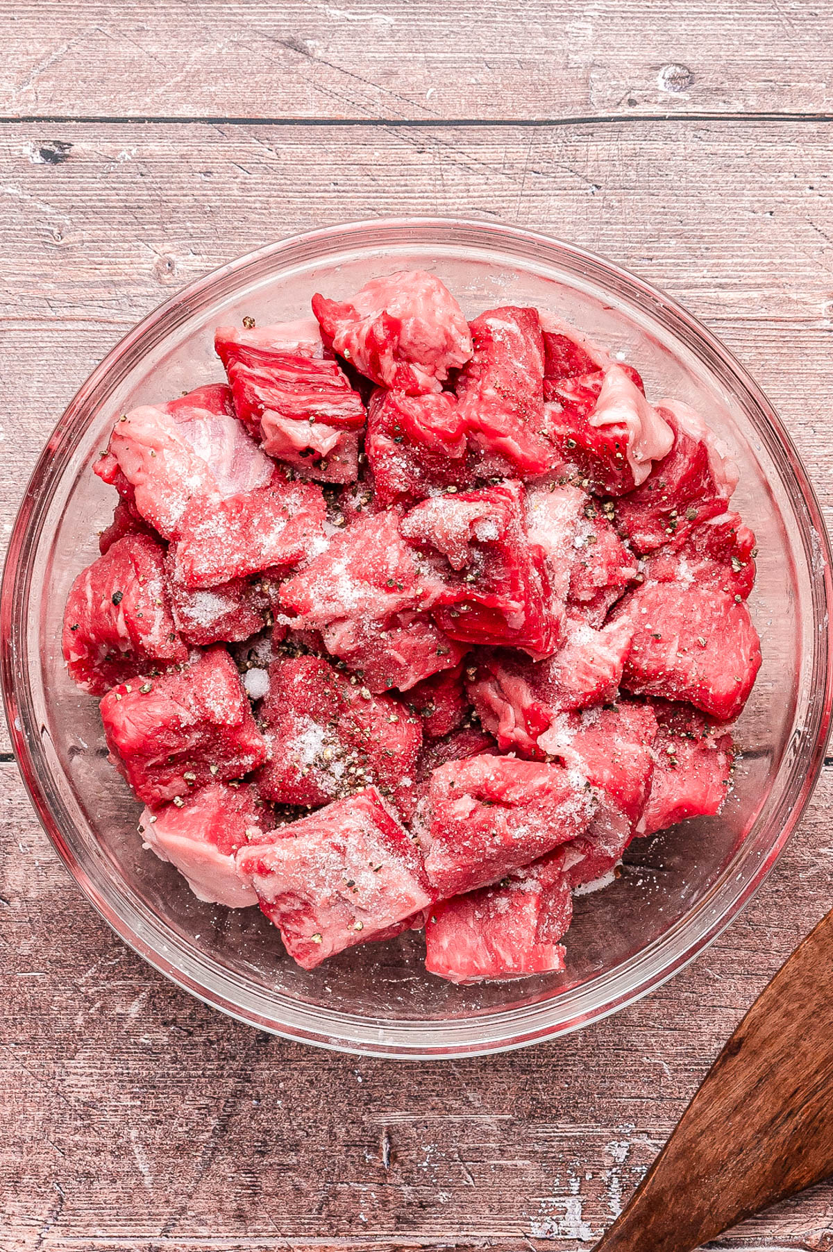 A glass bowl containing raw beef cubes seasoned with salt and pepper, placed on a wooden surface.