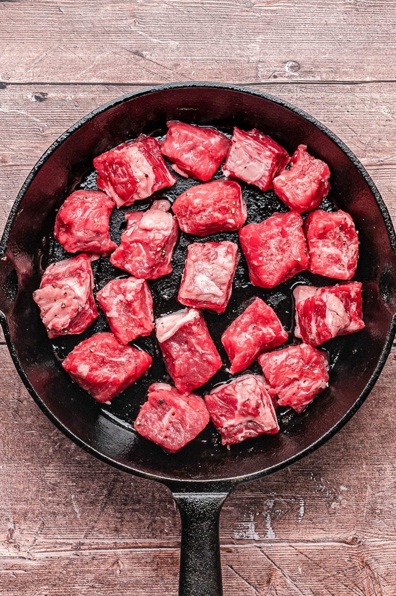 Raw beef cubes arranged in a single layer inside a black cast iron skillet on a wooden surface.