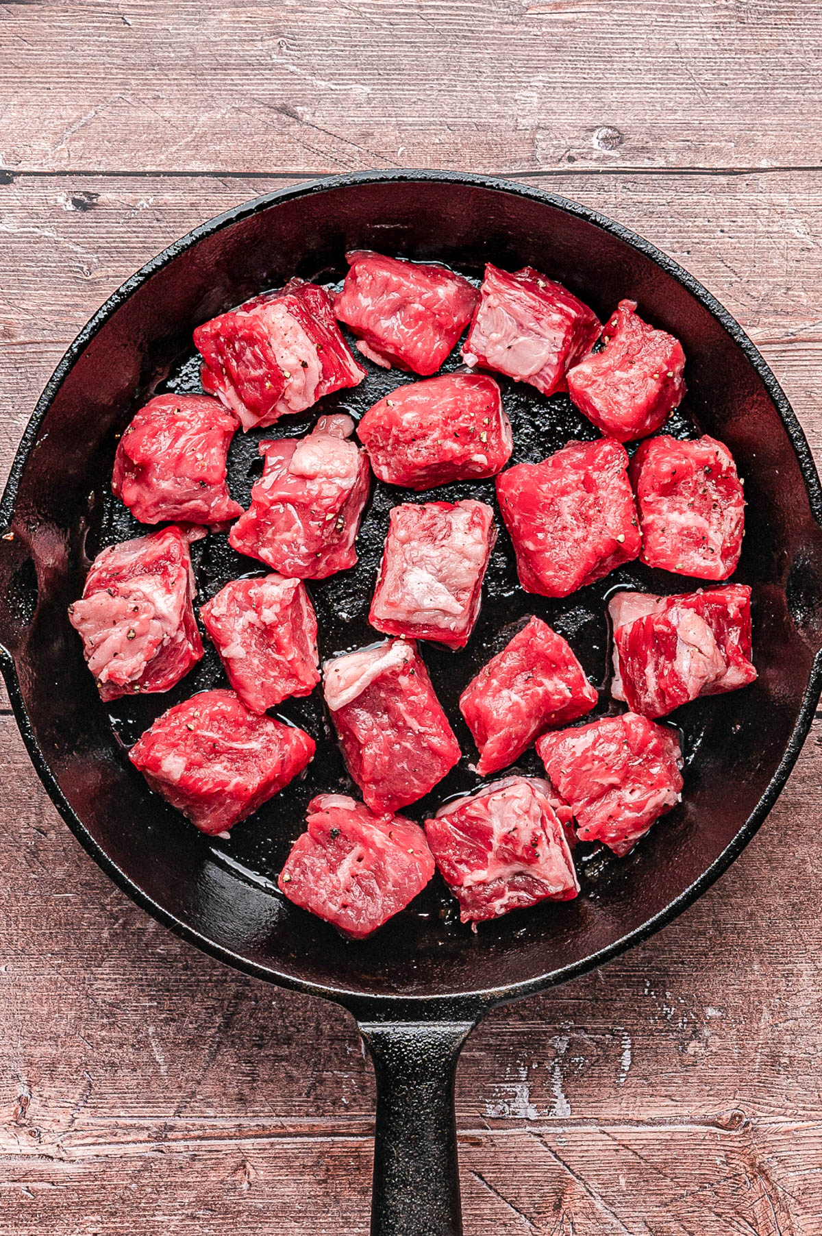 Raw beef cubes arranged in a single layer inside a black cast iron skillet on a wooden surface.