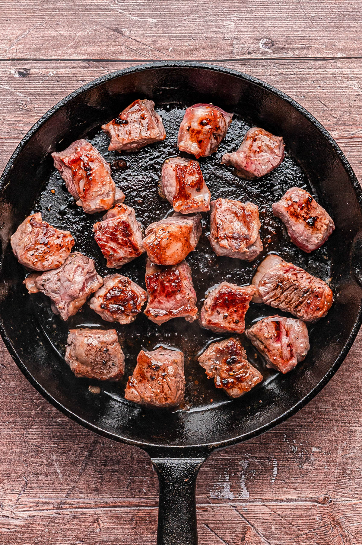 Chunks of beef are searing in a black cast iron skillet on a wooden surface.