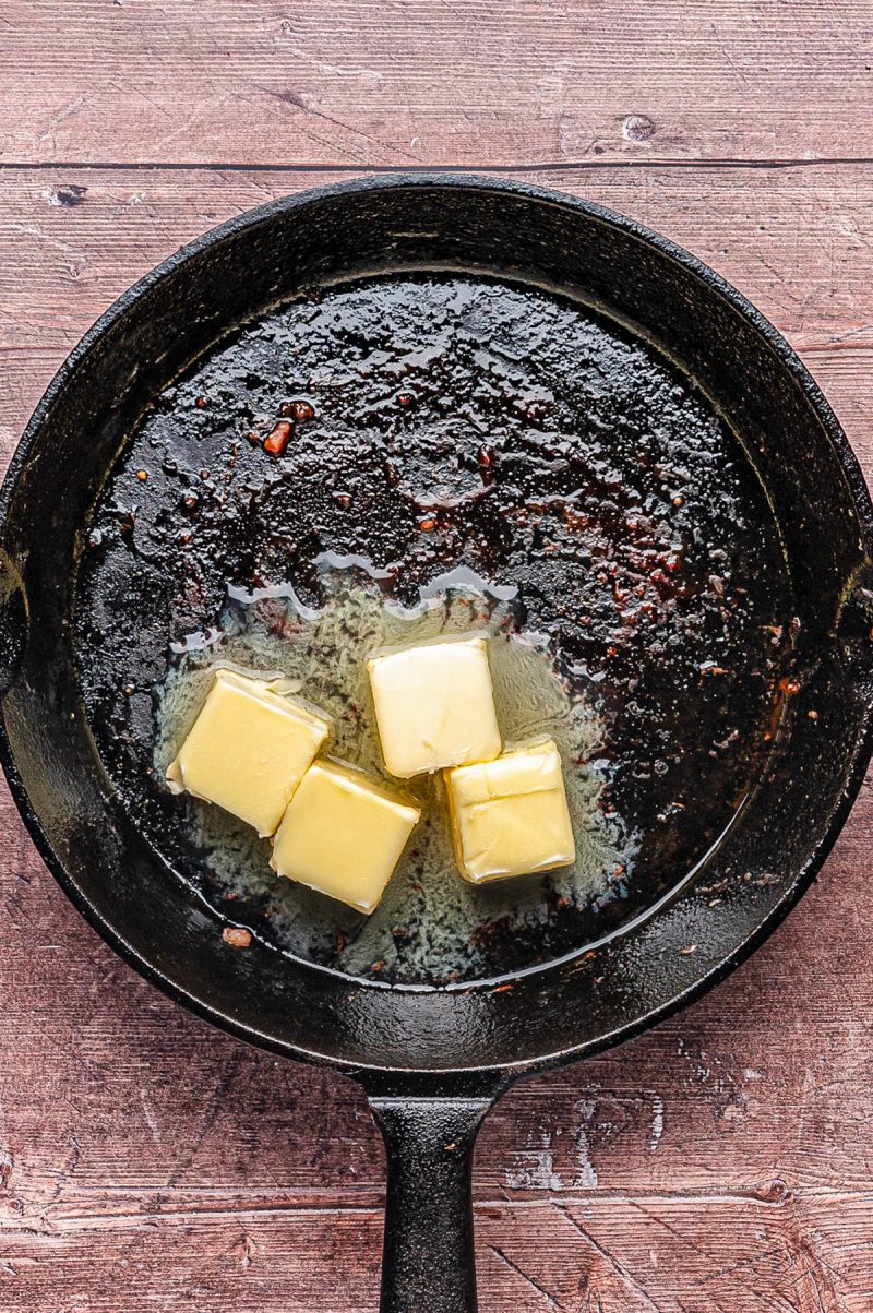 A black cast iron skillet with four cubes of butter melting in it, sitting on a wooden surface.