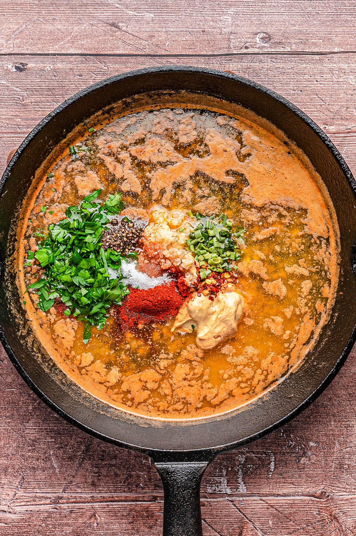 A cast iron skillet filled with a sauce mixture, topped with fresh chopped herbs, mustard, and various spices on a wooden surface.