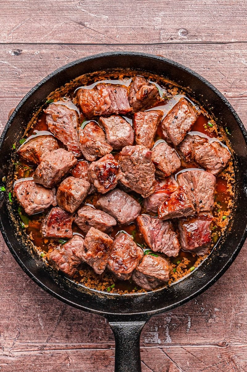 A black skillet filled with seared beef cubes in a brown sauce, resting on a rustic wooden surface.