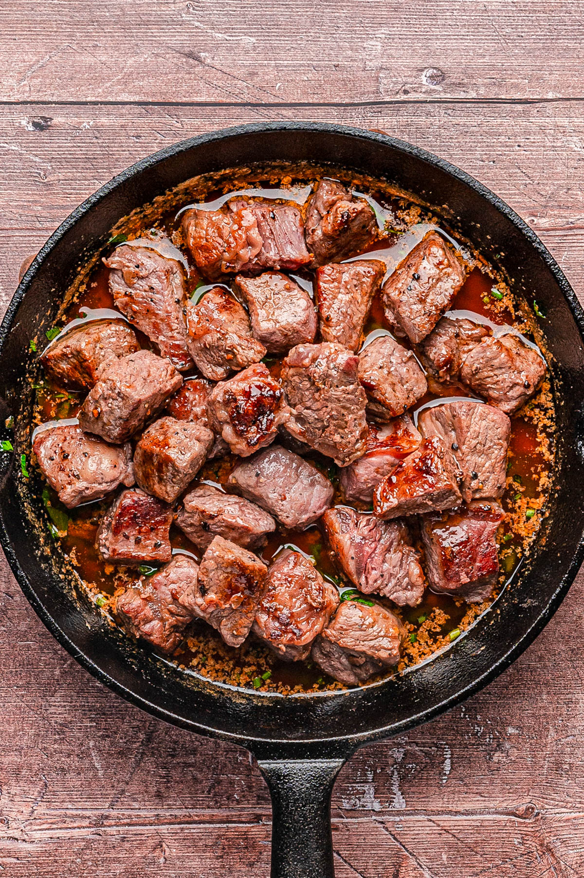 A black skillet filled with seared beef cubes in a brown sauce, resting on a rustic wooden surface.