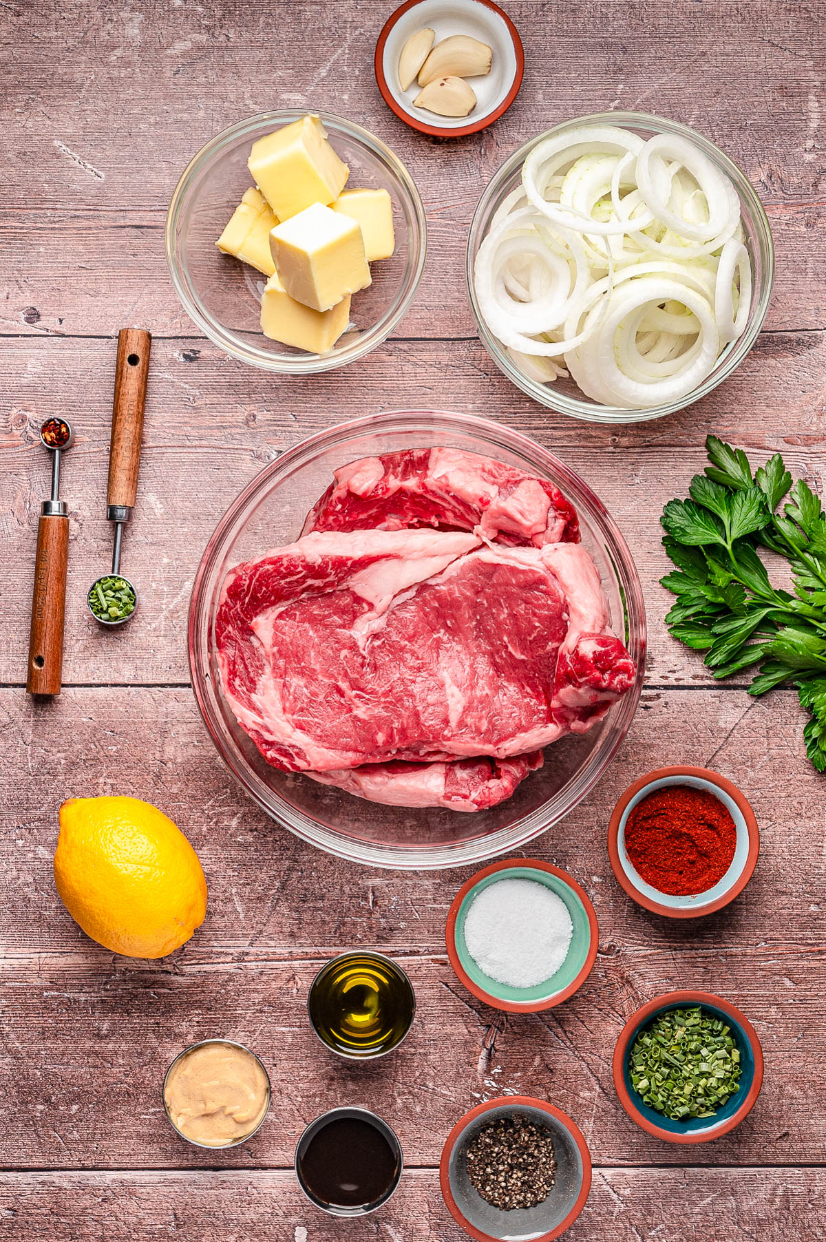 A variety of ingredients are arranged on a wooden surface, including raw steak, sliced onions, butter, spices, herbs, lemon, garlic, mustard, olive oil, and fresh parsley.