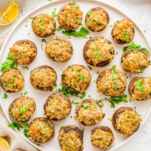 A plate of stuffed mushrooms garnished with chopped parsley, surrounded by lemon halves, fresh parsley, garlic, and breadcrumbs on a white surface.