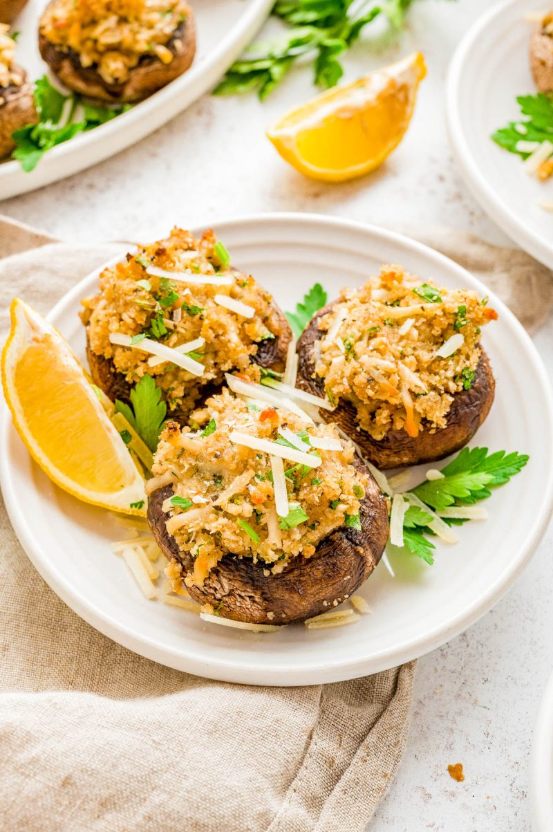 Three stuffed mushrooms topped with breadcrumbs and herbs are served on a white plate with shredded cheese, parsley, and lemon wedges.