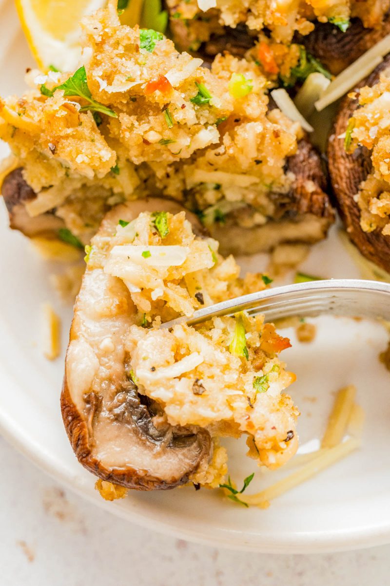 Close-up of a stuffed mushroom topped with breadcrumbs and herbs, with a fork cutting into it on a white plate.