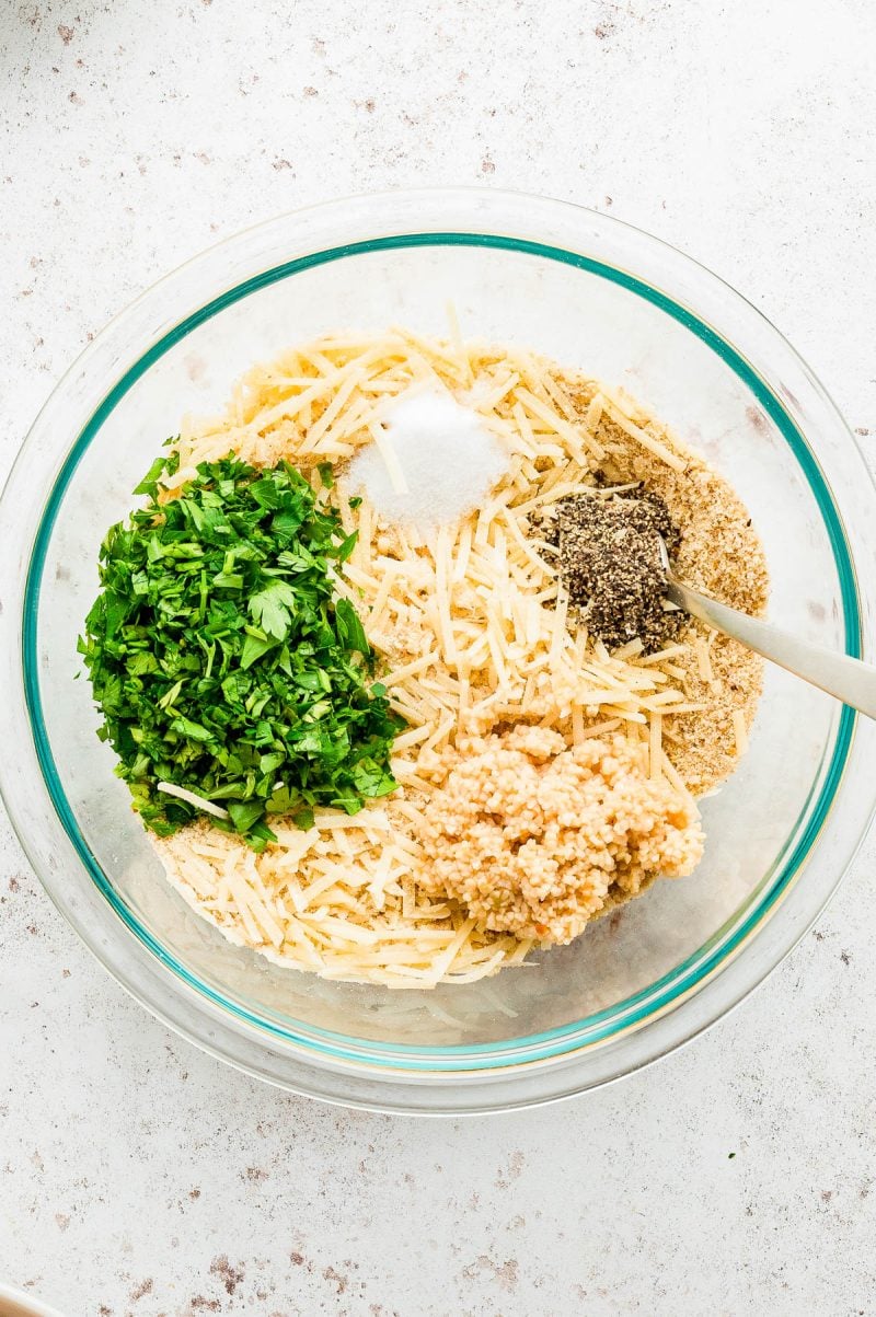 A glass bowl contains chopped parsley, grated cheese, breadcrumbs, minced garlic, ground black pepper, and salt, each in separate piles, with a spoon resting inside.