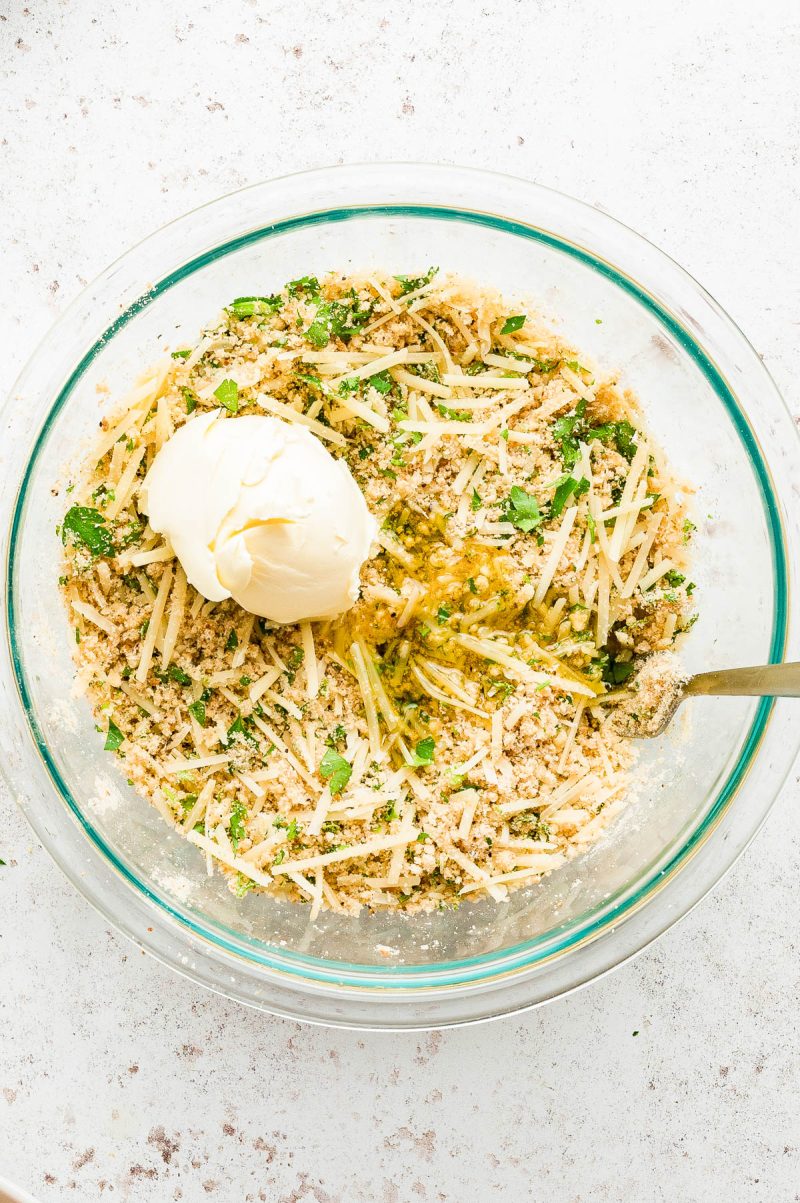 A glass bowl containing shredded cheese, breadcrumbs, chopped herbs, and a dollop of mayonnaise, with a metal spoon on the side.