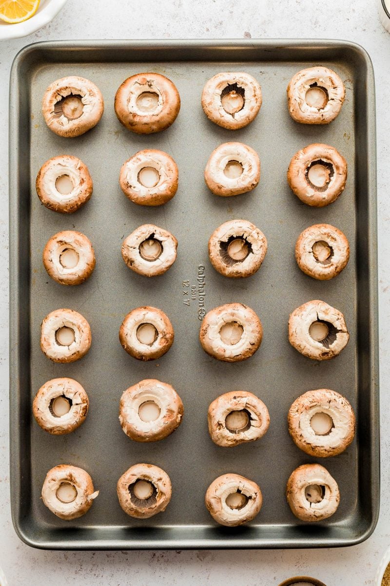 A baking sheet with 24 whole, hollowed-out mushrooms arranged in a grid pattern, ready for stuffing or roasting.