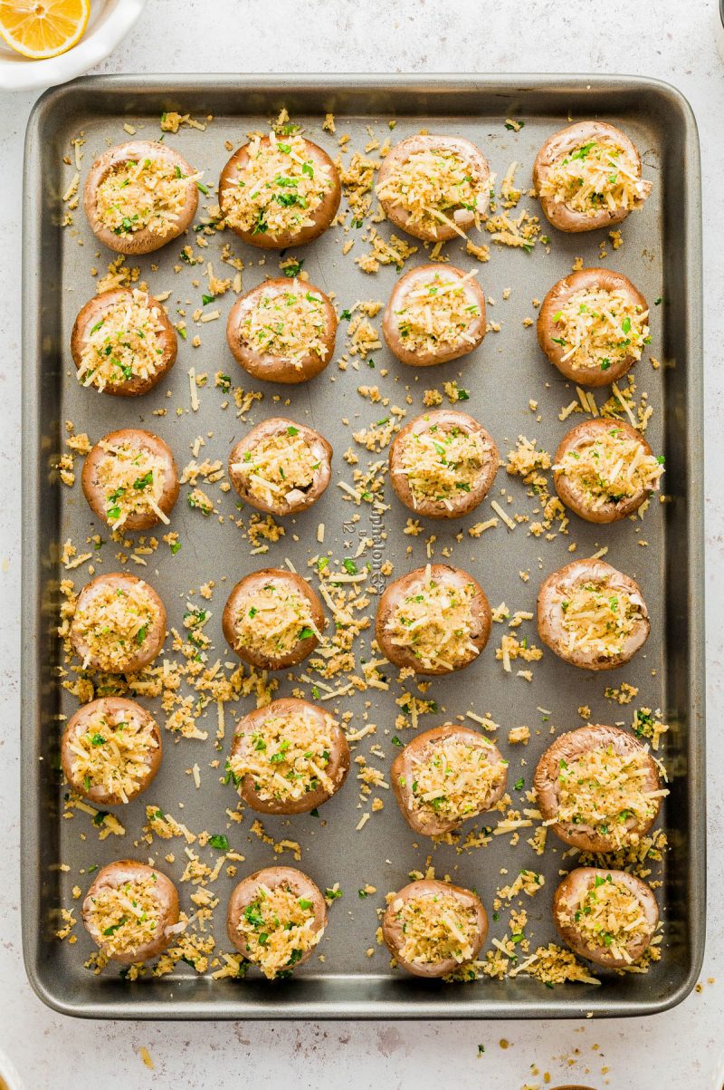 A baking sheet with rows of stuffed mushrooms topped with grated cheese and chopped herbs, ready to be baked.
