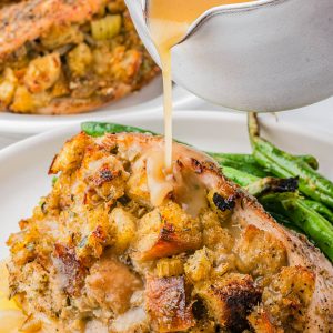 A close-up of gravy being poured over a baked, stuffing-topped chicken breast, served with green beans on a white plate.