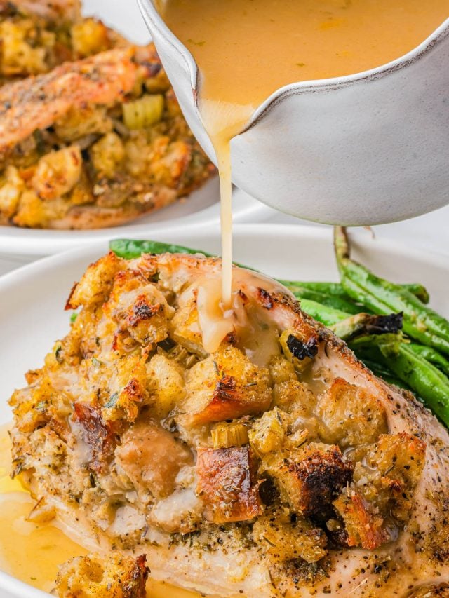 A close-up of gravy being poured over a baked, stuffing-topped chicken breast, served with green beans on a white plate.