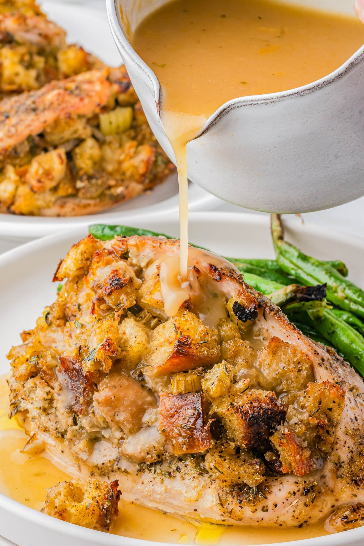 A close-up of gravy being poured over a baked, stuffing-topped chicken breast, served with green beans on a white plate.
