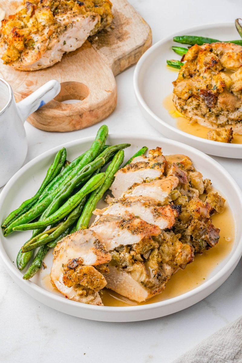 Sliced stuffed chicken breast with gravy, served with green beans on a white plate; additional servings and sauce in the background.
