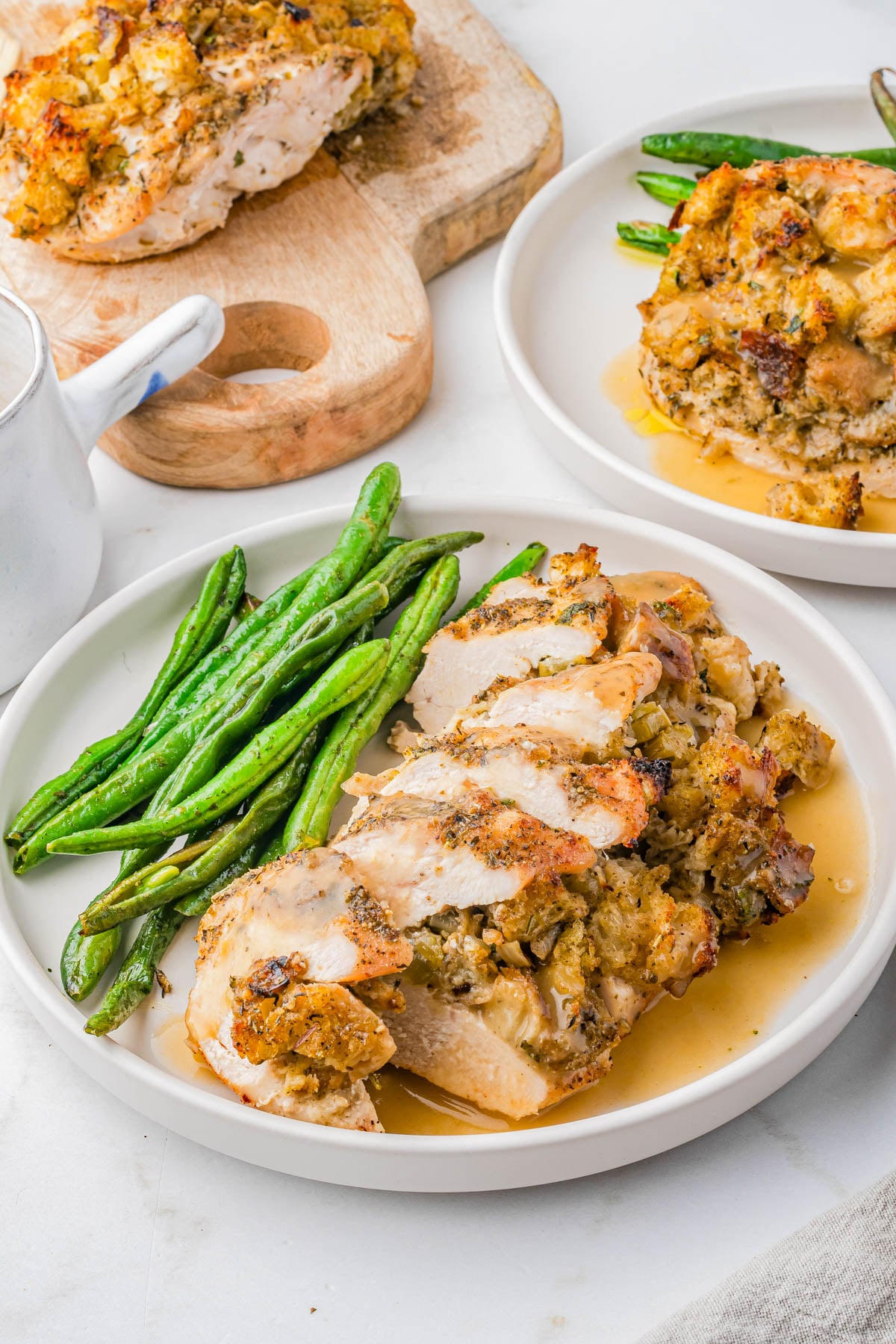 Sliced stuffed chicken breast with gravy, served with green beans on a white plate; additional servings and sauce in the background.