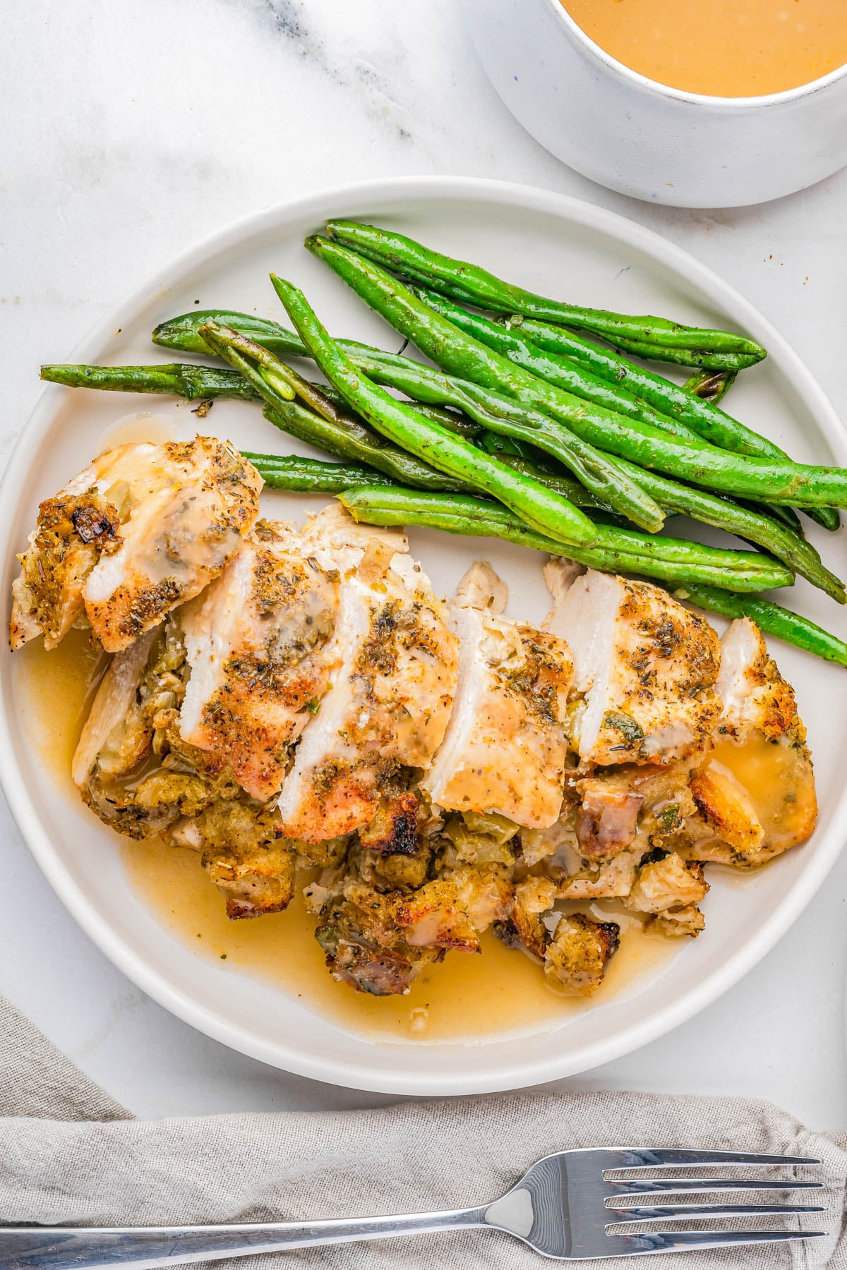 Sliced roasted chicken breast stuffed with dressing, served with green beans and gravy on a white plate.