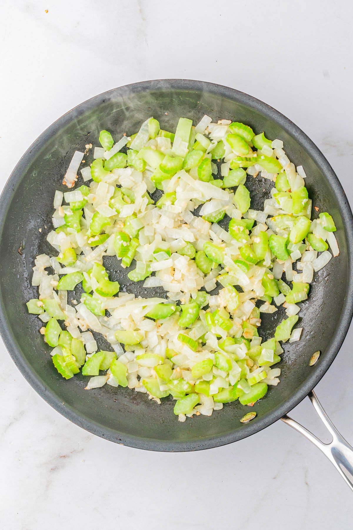Chopped onions and celery are being sautรฉed in a nonstick skillet on a white countertop.