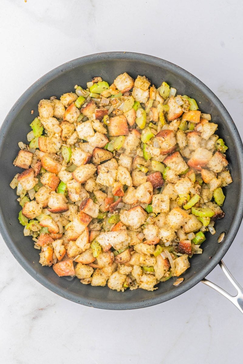 A skillet filled with bread cubes, chopped celery, and onions, mixed with herbs and seasonings, sits on a marble countertop.