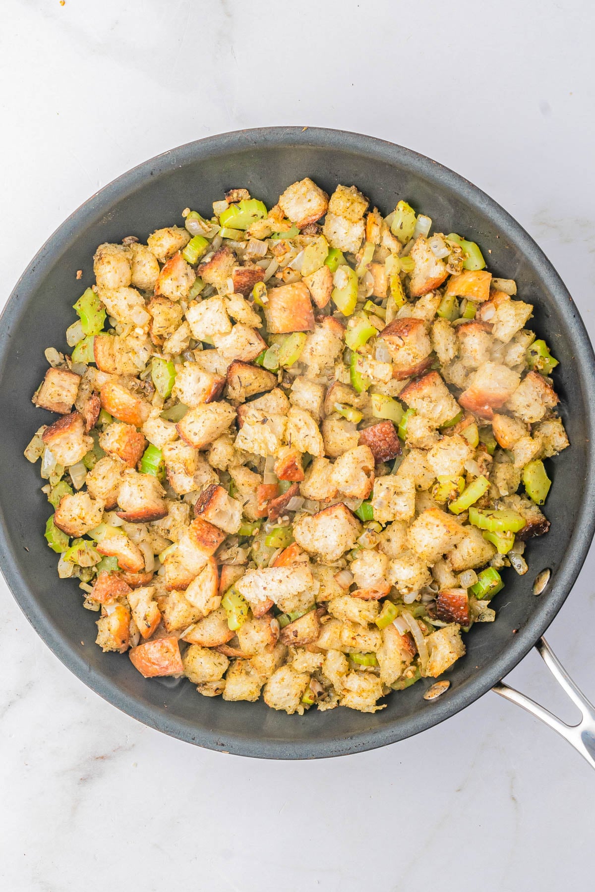 A skillet filled with bread cubes, chopped celery, and onions, mixed with herbs and seasonings, sits on a marble countertop.