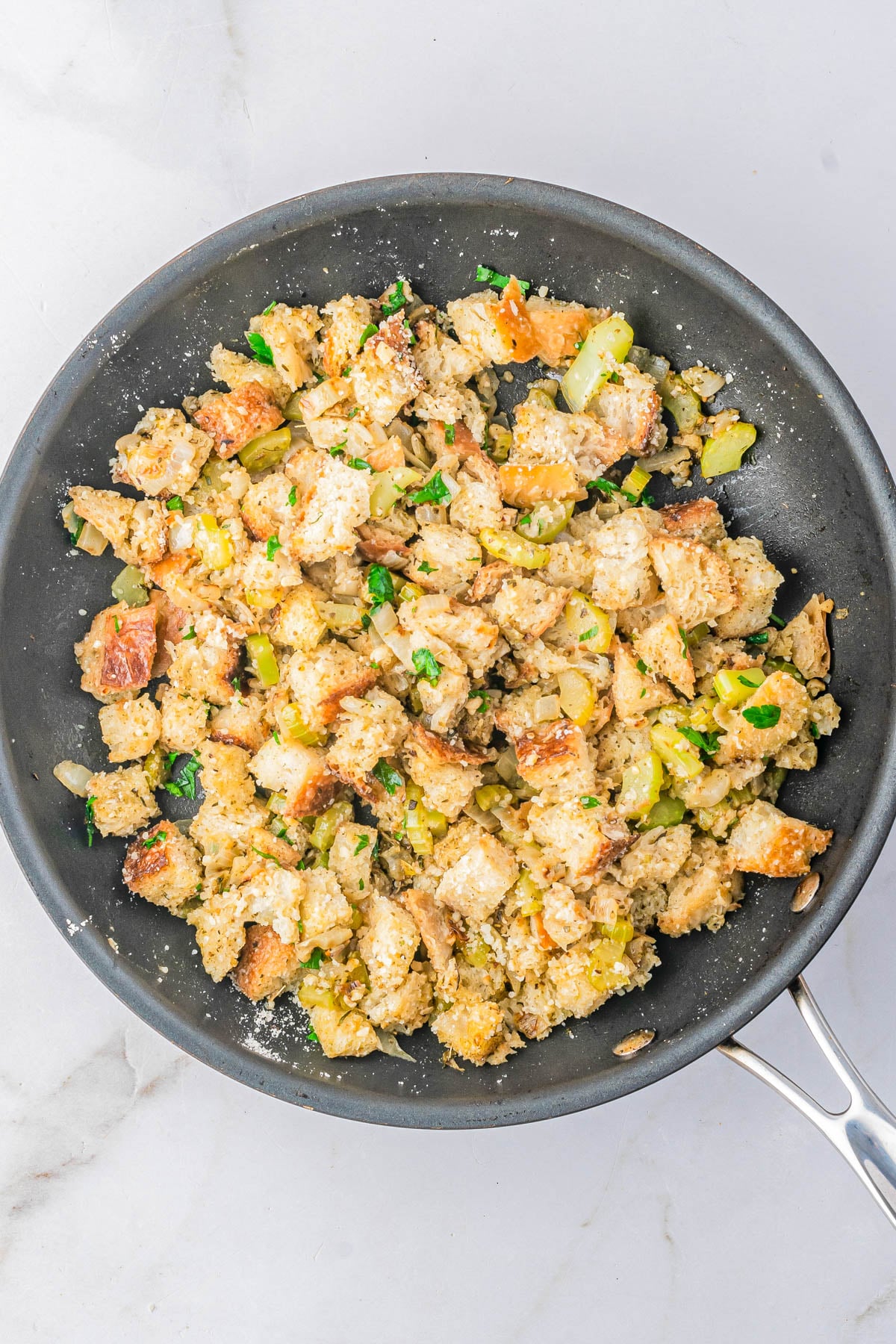 A skillet filled with bread stuffing mixed with celery, onions, herbs, and seasonings, sitting on a white countertop.