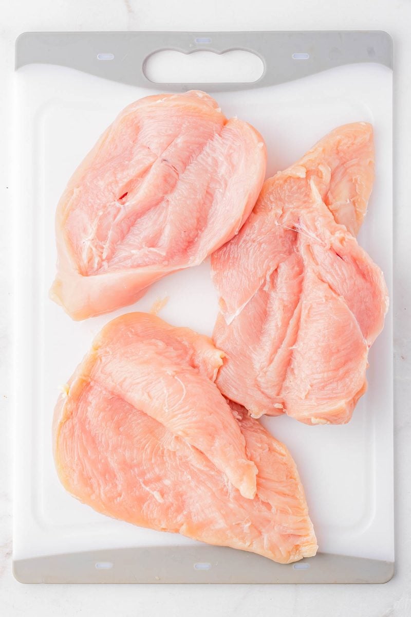 Three raw, boneless, skinless chicken breasts are placed on a white plastic cutting board.