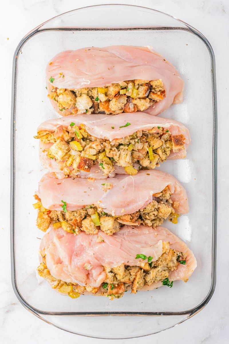Four raw chicken breasts in a glass baking dish, each sliced and stuffed with a bread and vegetable stuffing mixture.