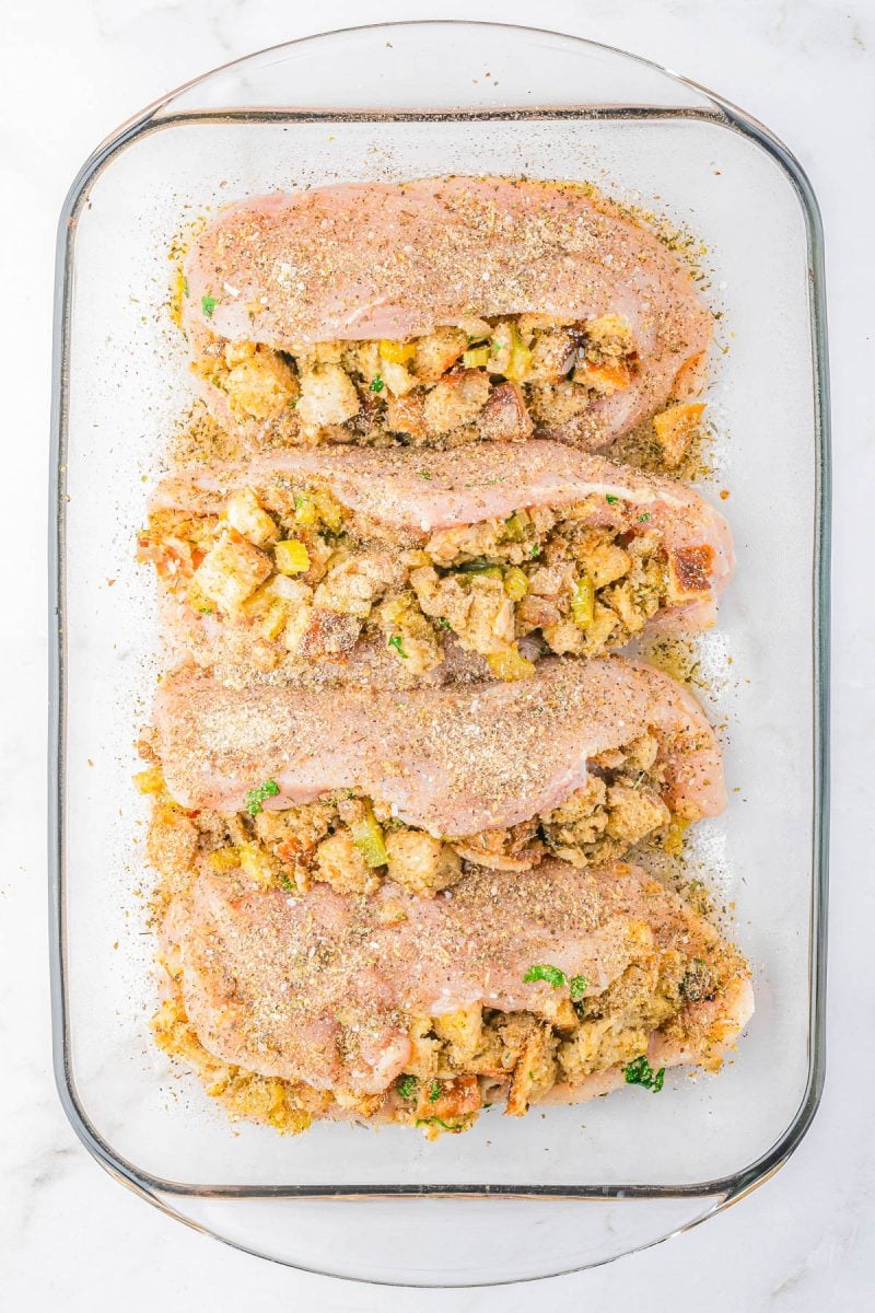 Four raw chicken breasts are slit and stuffed with bread stuffing, seasoned with spices, and arranged in a glass baking dish on a white background.