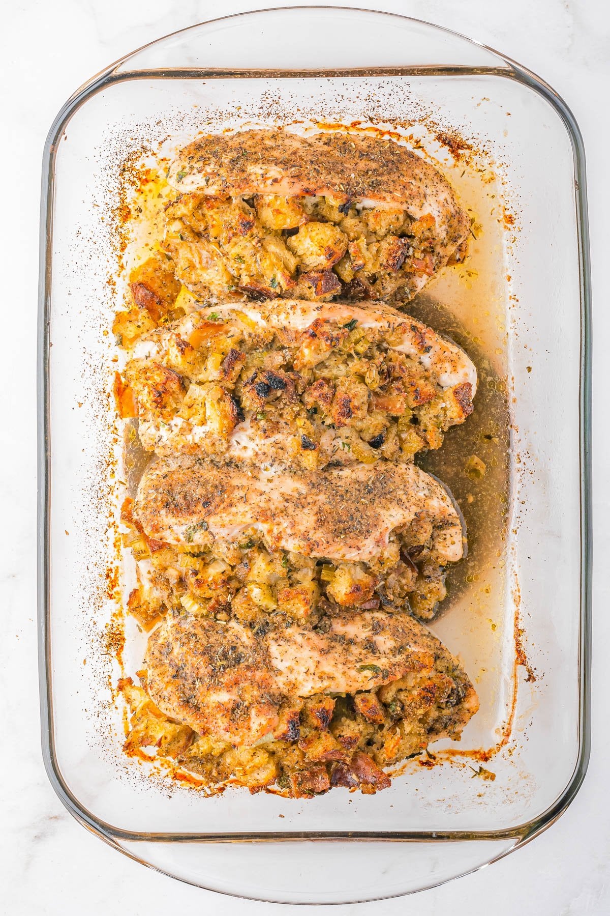 Four baked chicken breasts topped with stuffing in a glass baking dish on a white surface.